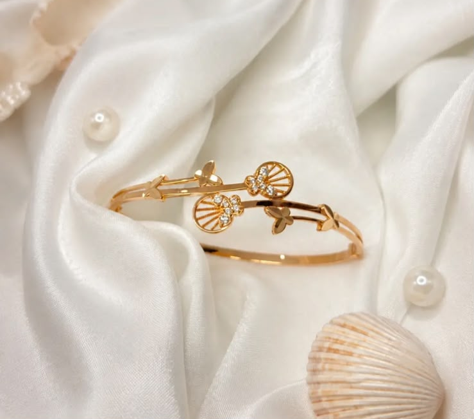 Gelang emas kaku (bangle) berdesain motif kerang laut dan kupu-kupu bertabur permata di atas kain satin putih.