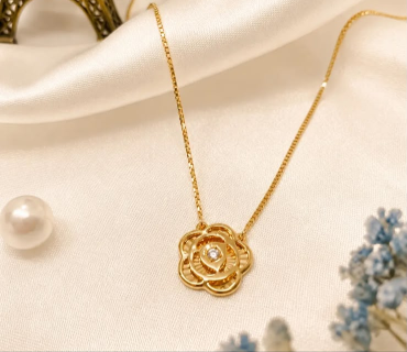 Kalung emas rantai halus dengan liontin berbentuk bunga mawar berhiaskan permata kecil di tengahnya di atas kain satin cream.
